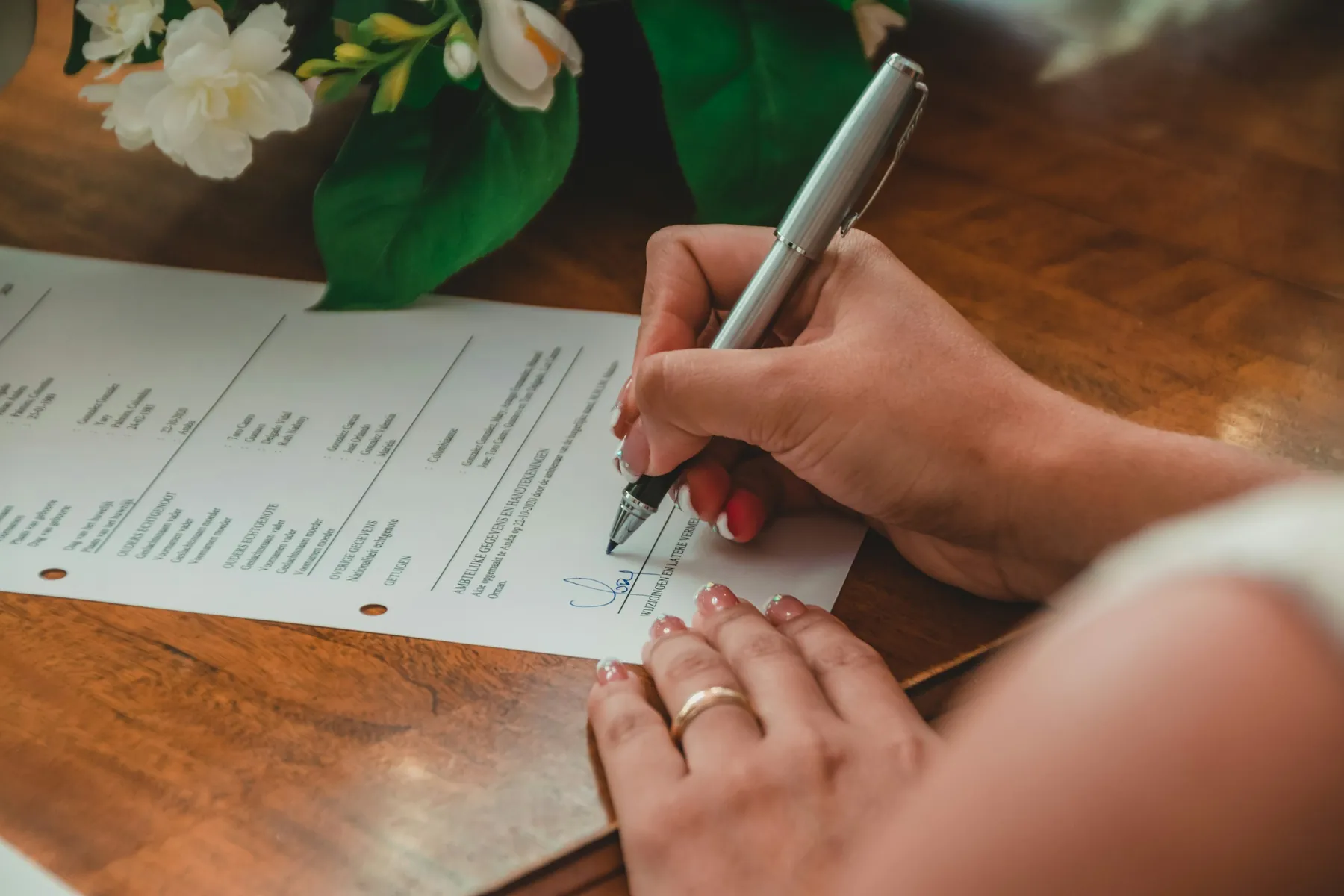 A hand signing a printed document with a silver pen on a wooden table, white flowers beside it.