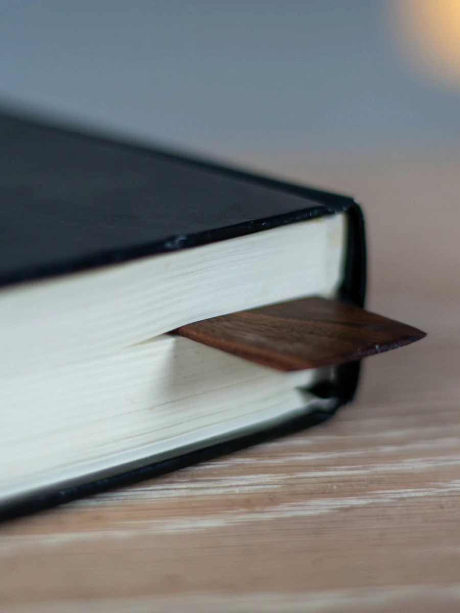A bound book closed on a wooden desk, a leather bookmark between its pages.