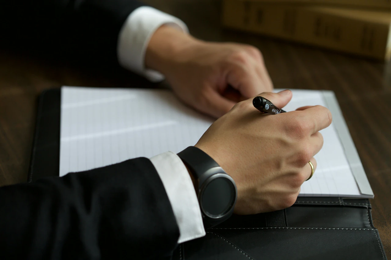A hand in a shirt cuff writing in a leather folio on a wooden desk.