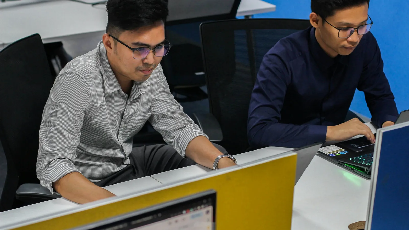 Two men in glasses working side by side at computers in an open office.